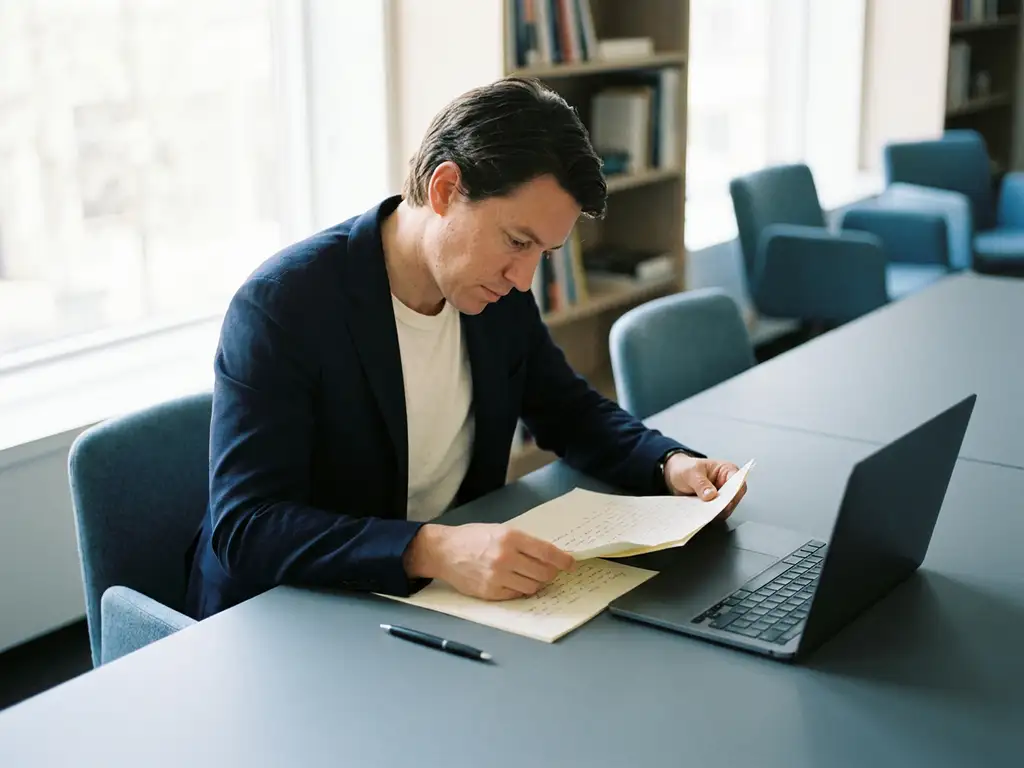 Job candidate reviewing handwritten notes at a modern interview desk with a closed laptop, bathed in soft natural window light.